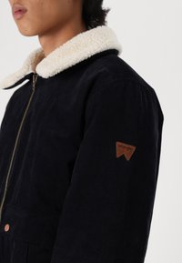 Granatowa kurtka z kordurowej tkaniny z kremowym kołnierzem z sherpy, z zapięciem na zamek błyskawiczny i brązową skórzaną naszywką z logo Wrangler na rękawie.