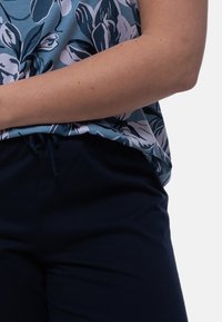 Nærbillede af en person, der bærer en blå blomsteret skjorte, der er stukket ned i mørkeblå bukser med snørelukning i taljen, mod en ensfarvet baggrund.