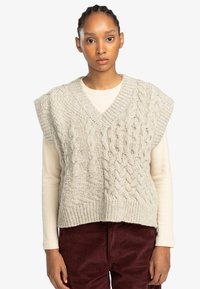Cable-knit sweater vest in light gray. Features a v-neck, short sleeves, and textured patterns. Worn over a cream long-sleeve top.