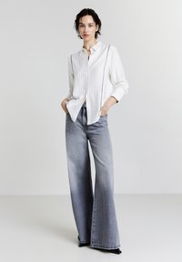 Donna con i capelli corti e bagnati, che indossa una camicia bianca con bottoni e righe verticali, e jeans a gamba larga di un blu sbiadito, con le mani nelle tasche.