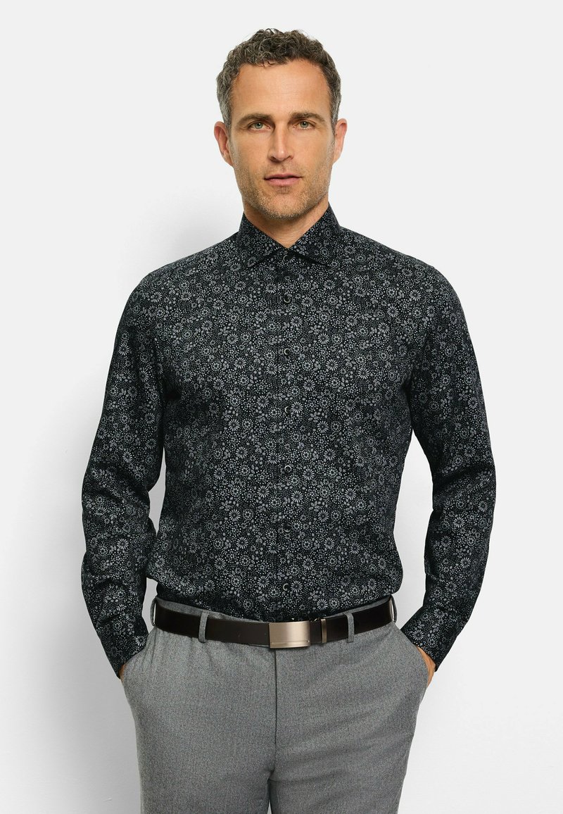 Schwarzes Langarmhemd mit einem weißen Blumenmuster, geknöpftem Kragen und einem glatten Stoff. Getragen mit grauen Hosen und einem Gürtel.