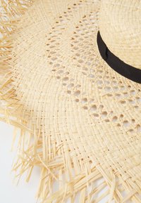 Leveled straw hat with a broad brim in natural beige color, featuring a textured weave and a black fabric band. The fringed edges add extra detail.