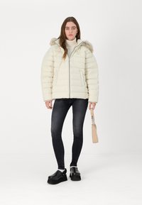 Doudoune écrue avec capuche en fourrure, dotée d'une fermeture éclair sur le devant et d'un motif matelassé horizontal, portée sur un jean skinny noir et des sabots.