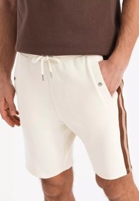 Shorts de couleur crème avec poches latérales, taille à cordon de serrage et accents de rayures brunes. Fabriqués en tissu doux avec une coupe décontractée.