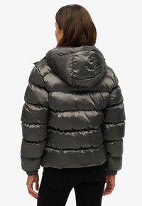 Kvinde iført en skinnende sort dunjakke med hætte, set bagfra, stående mod en ensfarvet hvid baggrund.