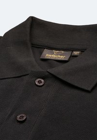 Schwarzes Poloshirt aus strukturiertem Stoff, mit klassischem Kragen und zwei dunklen Knöpfen. Das Etikett trägt die Aufschrift "Switcher made with respect."