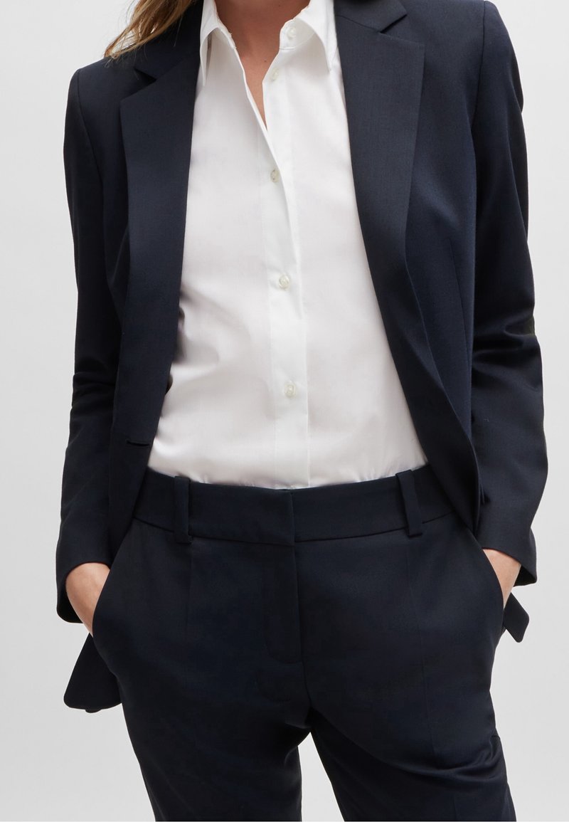 Femme portant une veste de costume noire ajustée et un pantalon avec une chemise blanche boutonnée, les mains dans les poches, sur un fond uni.