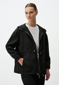 Zwarte windjack van zacht, lichtgewicht materiaal. Voorzien van een capuchon, elastische manchetten en een koord in de taille met gouden accenten.