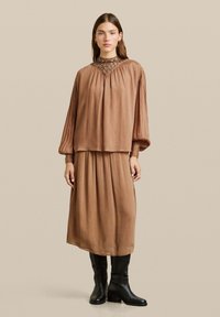 Vestito marrone plissettato con maniche lunghe a palloncino, tessuto testurizzato e scollatura impreziosita. Indossato con stivali neri con tacco al ginocchio.