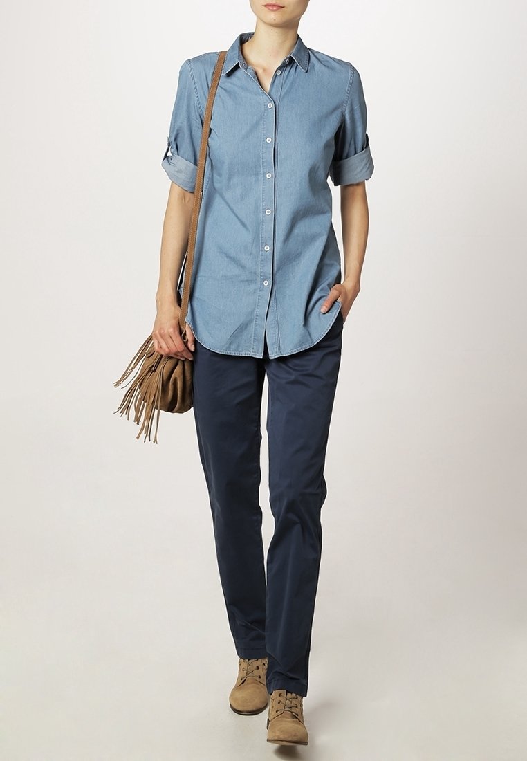 Femme portant une chemise bleu clair à manches retroussées, un pantalon bleu foncé, des chaussures cognac à lacets et un sac à épaule cognac à franges.