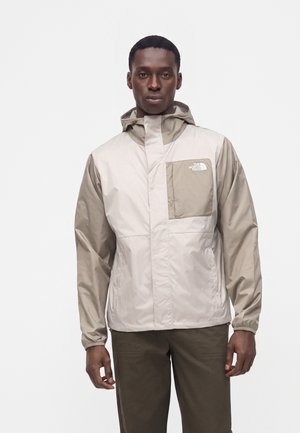 Homme portant une veste à capuche North Face beige et marron clair avec fermeture éclair et poche poitrine, debout sur fond blanc.