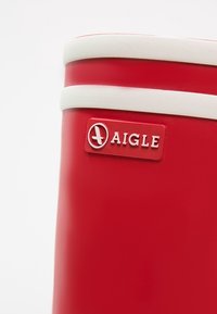 Botte en caoutchouc rouge avec un liseré blanc présentant un logo Aigle embossé près du bord supérieur.