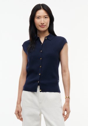 Vrouw met lang zwart haar, gekleed in een marineblauw mouwloos knoophemd en witte broek tegen een effen witte achtergrond.