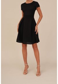 Robe noire ajustée évasée avec manches courtes, tissu texturé, jupe plissée et encolure ronde. Portée avec des talons métalliques à bride cheville.