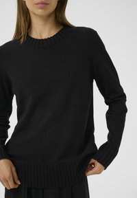 Frau, die einen schwarzen Strickpullover mit geripptem Rundhalsausschnitt, Bündchen und Saum trägt und mit einer Hand den unteren Rand vor einem weißen Hintergrund anpasst.