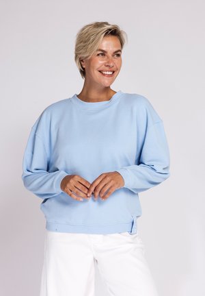 Smiling woman with short blonde hair wearing a light blue sweatshirt and white pants, standing against a plain light background.