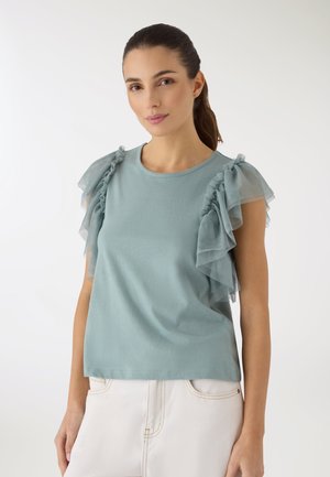 Donna che indossa una maglia azzurro chiaro con maniche arricciate e pantaloni bianchi, in piedi davanti a uno sfondo bianco semplice.