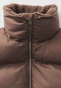 Doudoune marron rembourrée avec un col court et duveteux. Possède une fermeture éclair métallique et une texture de tissu lisse. Design minimaliste, sans motifs visibles.