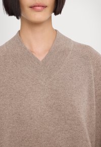 Femme portant un pull en laine beige à col en V avec un col côtelé et des détails aux épaules, visible du menton à la poitrine sur un fond blanc.