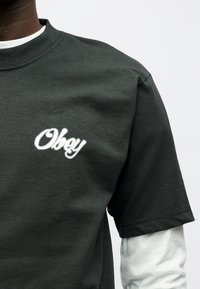 Άτομο που φοράει σκούρο πράσινο κοντομάνικο πουκάμισο πάνω από λευκό μακρυμάνικο πουκάμισο, με τη λέξη «Obey» γραμμένη με λευκά γράμματα στο στήθος.