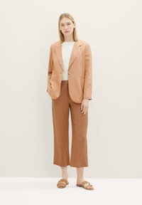 Blazer en lin marron clair avec des revers à cran, associé à un pantalon large marron foncé et des sandales à glissière marron. Design simple et texturé.