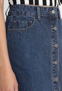 Falda de mezclilla en azul oscuro, con una cintura alta, cierre de botones en el frente y bolsillos laterales. La textura es suave con un ligero estiramiento.