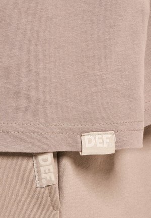 Beige bomull t-skjorte med en myk tekstur, med rett nederkant og to synlige etiketter sydd nederst, merket "DEF."