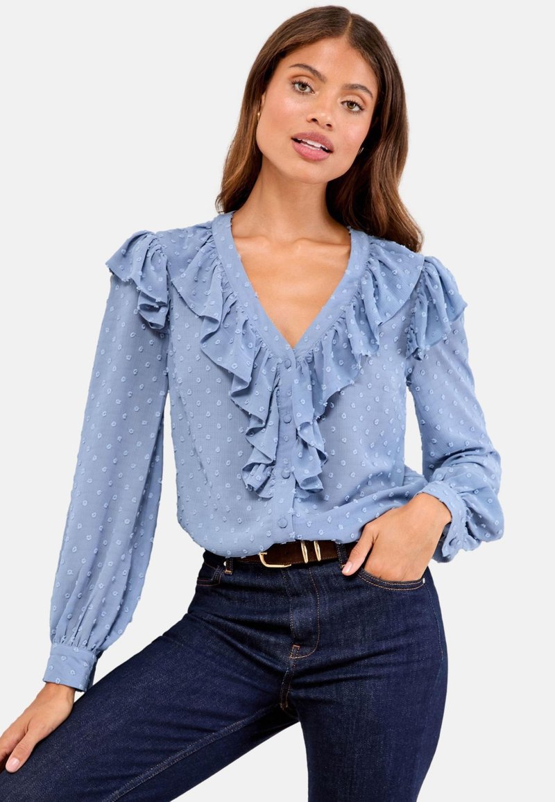 Woman wearing a light blue long-sleeve blouse with textured dots and ruffled V-neck paired with dark blue jeans and a black belt.