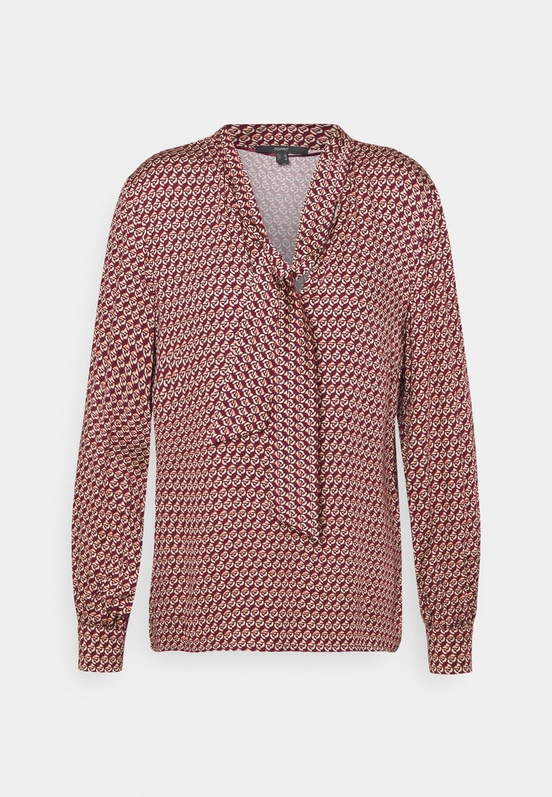 Blouse rouge à manches longues, avec un col noué et un motif répétitif de petites formes dans des tons crème et rouge foncé. Texture de tissu douce.
