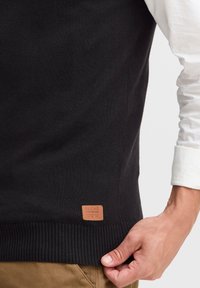 Svart stickad väst med ribbad nederkant, utrustad med en tanfärgad läderetikett med texten "BLEND". Mjuk textur och åtsittande design.