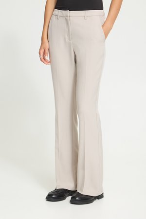 Pantalon beige à jambes droites porté avec des chaussures noires et un haut noir, une main dans la poche et l’autre reposant sur le côté.