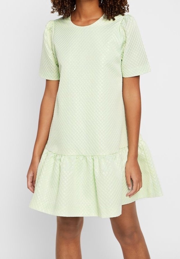 Jeune femme portant une robe courte à manches courtes texturée vert clair avec une taille basse et un ourlet froncé, sur un fond uni.