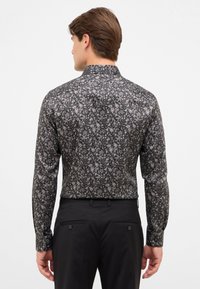 Mann, der sich abwendet, trägt ein langärmliges schwarz-weißes Blumenhemd, das in schwarze Hosen gesteckt ist, vor einem schlichten Hintergrund.