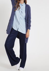 Dlouhý tmavě modrý pletený cardigan přes světle modrou košili s knoflíky, doplněný širokými tmavě modrými kalhotami a bílými teniskami. Texturovaná tkanina, volný střih.