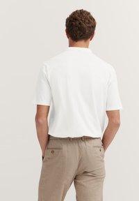 Weißes Polo-Shirt mit kurzen Ärmeln und lockerer Passform, kombiniert mit hellbraunen Leinenhosen, die eine Gesäßtasche und Knopfdetails aufweisen.