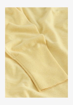 Gaiši dzeltena adīta auduma ar mīkstu tekstūru, kurai ir ribota aproce. Visā materiālā ir redzami maigi locījumi un kaskādes.