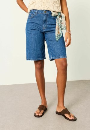 Személy zászlós, mintás sállal kötött övet visel kék térd alá érő farmer rövidnadrághoz, krém színű kötött felsőt, gyöngyös karkötőt és barna szandált.