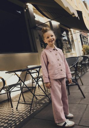 Ung pige i lyserød fløjlsjakke og bukser smilende og stående ved siden af sorte metalstole og borde på et mønstret fortov uden for en café.