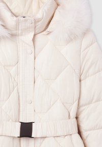 Giacca puffer trapuntata in crema con cappuccio in pelliccia sintetica, bottoni a scatto e una vita nera con cintura. Presenta una cucitura diagonale e una texture liscia.
