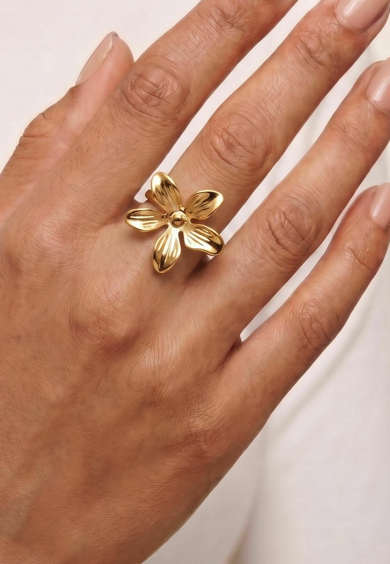 Main à la peau brun moyen portant une grosse bague en or en forme de fleur à quatre pétales au doigt du milieu.