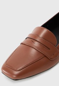 Mocassin en cuir marron avec un bout carré, présentant une sangle horizontale sur le dessus pour le détail et une texture lisse.
