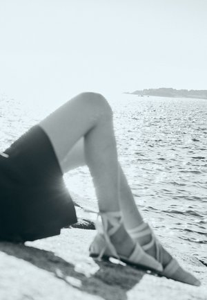Osoba koja se odmara kraj vode, nosi crne kratke hlače i šiljate, remenaste cipele, noge ispružene prema moru i vidljiva udaljena obala.