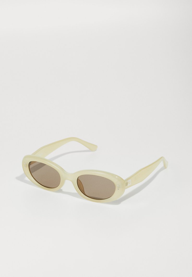 Gafas de sol ovaladas con un marco translúcido de color amarillo pálido y lentes marrones. Presenta una textura suave y sutiles acentos metálicos en los laterales.