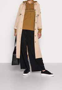 Trench coat beige con bottoni, felpa marrone con testo nero, pantaloni larghi neri e sneakers nere con suola bianca.
