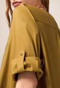 Senfgelbes Hemd mit aufgerollten Ärmeln, glatter Textur, Knopfdetails an den Manschetten und einer lockeren Passform. Subtile Glanz und breites Kragen-Design.