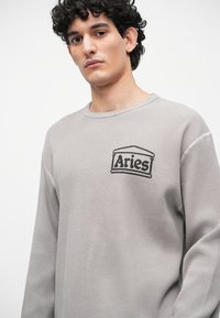 Szary sweter z dzianiny w zależności od warkocza, z czarnym haftowanym logo "Aries" na klatce piersiowej oraz okrągłym dekoltem i długimi rękawami.