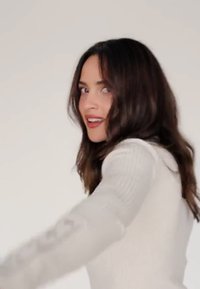 Una donna con lunghi capelli scuri, che indossa un maglione a coste grigio chiaro, si gira leggermente, assumendo una posa rilassata su uno sfondo semplice.