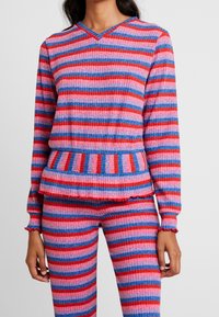 Wielokolorowy sweter z dzianiny i dopasowane spodnie. Posiada czerwone, różowe i niebieskie poziome paski z prążkowaną fakturą i falbankowymi mankietami.