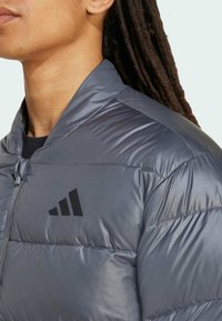 Profil osoby ubranej w szary puchowy kurtkę z czarnym logo na piersi. Osoba ma długie, ciemne, stylizowane włosy.