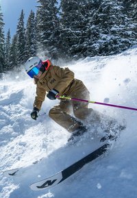Escena de esquí con una persona en un traje de esquí beige con acentos negros, casco blanco y gafas de sol espejadas azules, esquiando a través de nieve polvo.